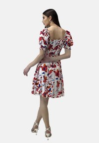 Vestido floral con mangas abullonadas, que presenta un escote cuadrado, patrones rojos y azules sobre una tela blanca, y una falda acampanada. Sandalias de tacón blancas.