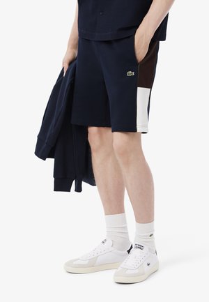Shorts de sport navy avec des panneaux latéraux blancs et marron, en tissu mélangé coton, avec des poches, associés à des baskets blanches et des chaussettes montantes.