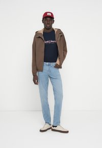 Καφέ τζάκετ με μπεζ επένδυση, μπλε μπλουζάκι με λογότυπο, ανοιχτό μπλε τζιν και κρεμ αθλητικά παπούτσια με μαύρες λεπτομέρειες, σε συνδυασμό με κόκκινο καπέλο.