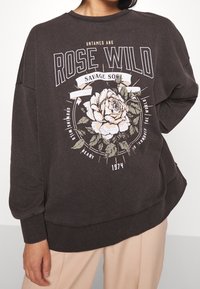 Svetle šedá mikina s volným střihem, s květinovou grafikou v bílé a zelené barvě, s nápisem "Rose Wild" a "Savage Soul."