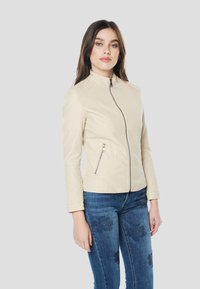 Beige nepleren jas met een hoge kraag, diagonale ritssluiting en doorgestikte accenten op de schouders en mouwen. Slim-fit ontwerp, met een gestructureerde vorm.