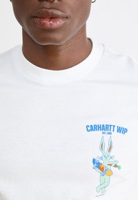T-shirt bianca in cotone con una grafica blu e verde che rappresenta un coniglio con uno skateboard e una carota, insieme al testo "CARHARTT WIP EST. 1989".