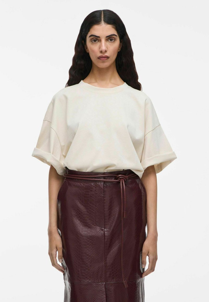 Beige oversized T-shirt met korte mouwen, gedragen met een bordeauxrode leren rok met slangenprint en een strikdetail in de taille.