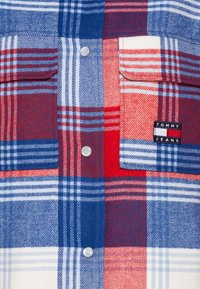Camicia in flanella a quadri rossi, bianchi e blu, con due tasche sul petto, bottoni a pressione argentati e un piccolo etichetta con il logo Tommy Jeans.