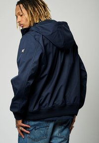 Veste à capuche navy avec fermeture éclair, poignets élastiques et ourlet côtelé. Présente une étiquette de logo sur la manche et une texture de tissu lisse.