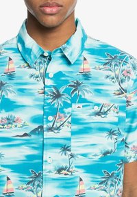 Lyhythihainen nappipaita turkoosilla taustalla, jossa on trooppinen kuvio palmuista ja purjeveneistä; mukana rintatasku.