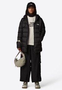 Svart pufferjakke, sweatshirt med hvitt logo, svarte vide bukser, grå skulderveske og beige joggesko, med en svart caps som fullfører utseendet.
