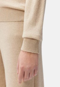 Beige stickad tröja med ribbad mudd och en slät yta. En hand är delvis synlig, vilande mot ett matchande beige plagg.