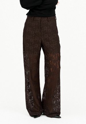 Pantalon large en dentelle marron à taille haute, porté par-dessus des chaussures noires pointues, associé à un haut noir à manches longues.