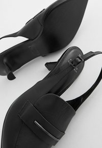 Svarta läderslingback-halkningar med rund tå, liten blockklack och dekorativ rem. Texturerad sula för bra grepp.