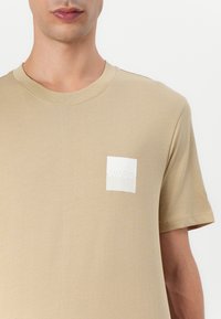 T-shirt en coton beige avec un col rond et des manches courtes, arborant un petit logo carré blanc sur la zone gauche de la poitrine. Texture lisse.