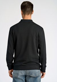 Pullover lungo nero a maniche lunghe con colletto e polsini a coste, caratterizzato da una texture liscia e una vestibilità comoda, abbinato a jeans azzurri chiari.