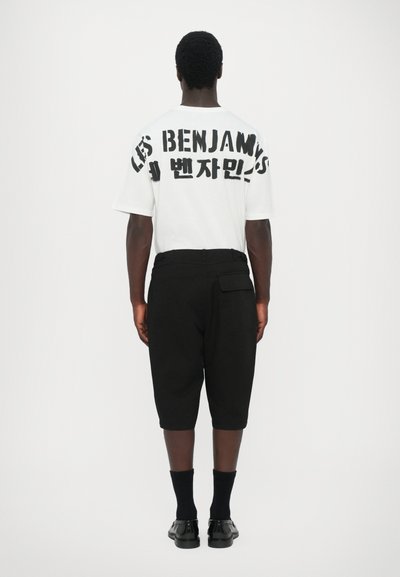 T-shirt blanc avec un texte noir surdimensionné au dos, associé à un pantalon noir cropped. Des chaussettes et des chaussures noires complètent la tenue.