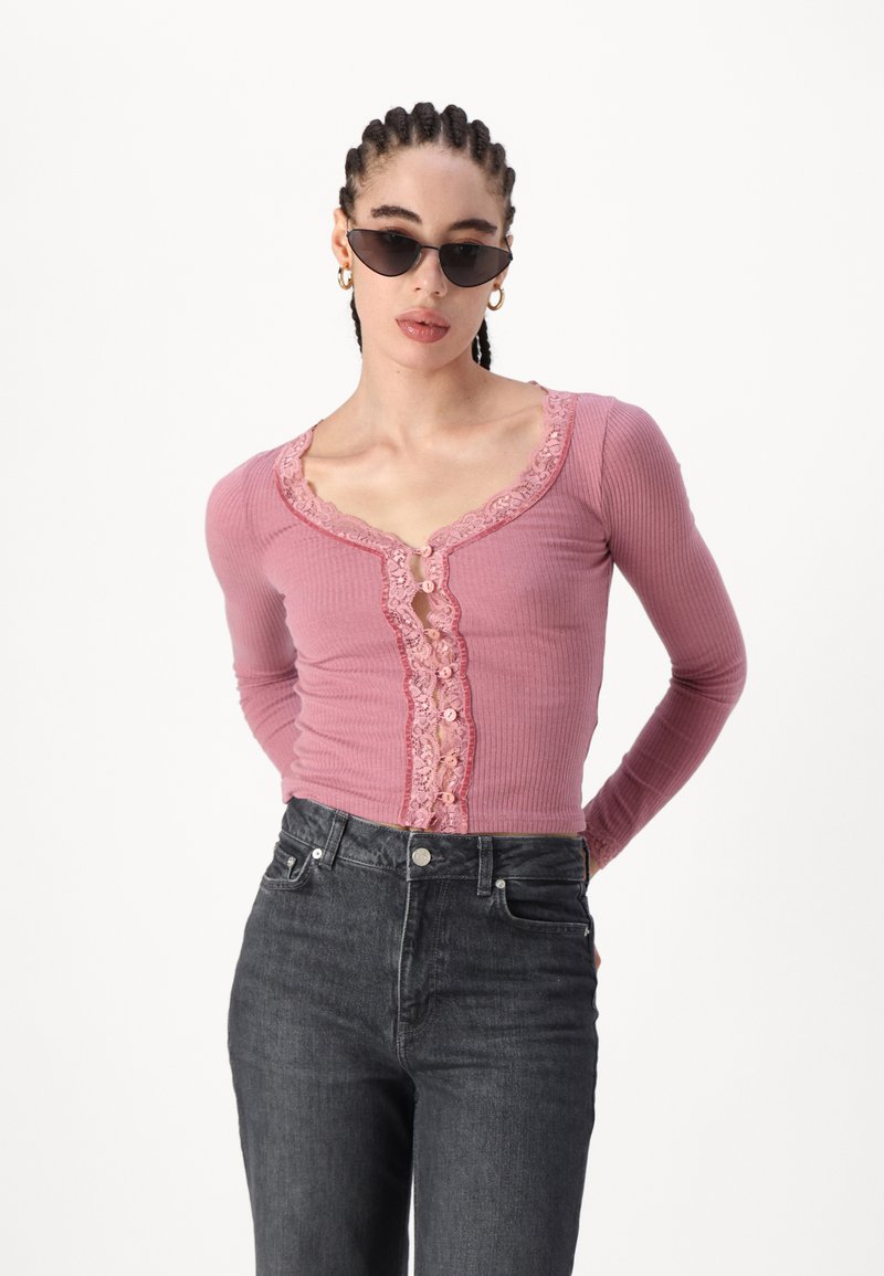 Haut à manches longues en rose côtelé avec finition en dentelle et détail de boutons, associé à un jean foncé. Présente un décolleté arrondi et une coupe ajustée.