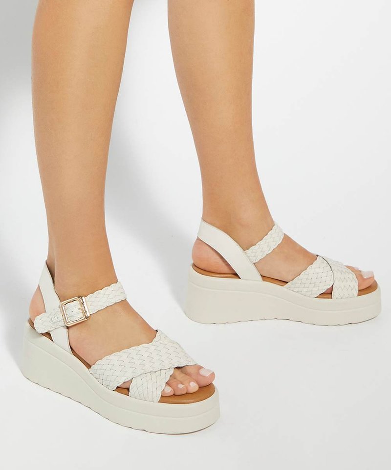 Pieds portant des sandales compensées blanches tressées avec des brides à la cheville et des boucles dorées sur un fond blanc uni.