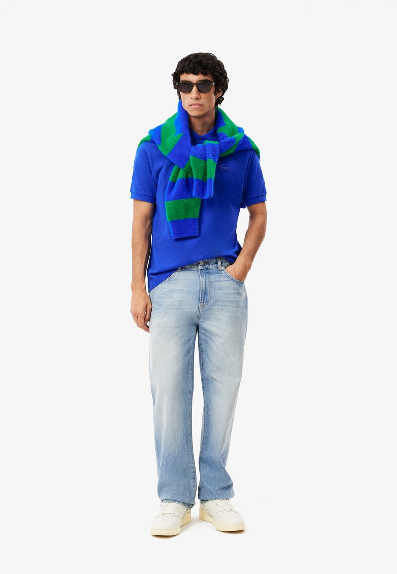 Blaues Poloshirt, hellblaue Jeans, weiße Sneakers und ein grün-blau gestreifter Schal um den Hals drapiert; Sonnenbrille rundet das Outfit ab.