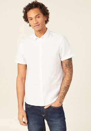 Camisa blanca de manga corta y botones, hecha de lino ligero, con cuello clásico y puños remangados, combinada con unos vaqueros de mezclilla azul oscuro.