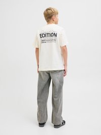 Kremowa bawełniana koszulka z czarnym nadrukiem na plecach, zestawiona z luźnymi szarymi dżinsami i czarnymi sandałami.