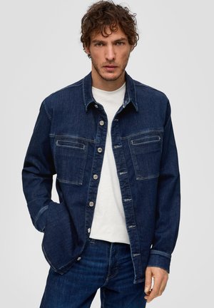 Dunkelblaue Jeansjacke mit Knopfverschluss, zwei Brusttaschen und kontrastierenden Nähten. Getragen über einem weißen T-Shirt.