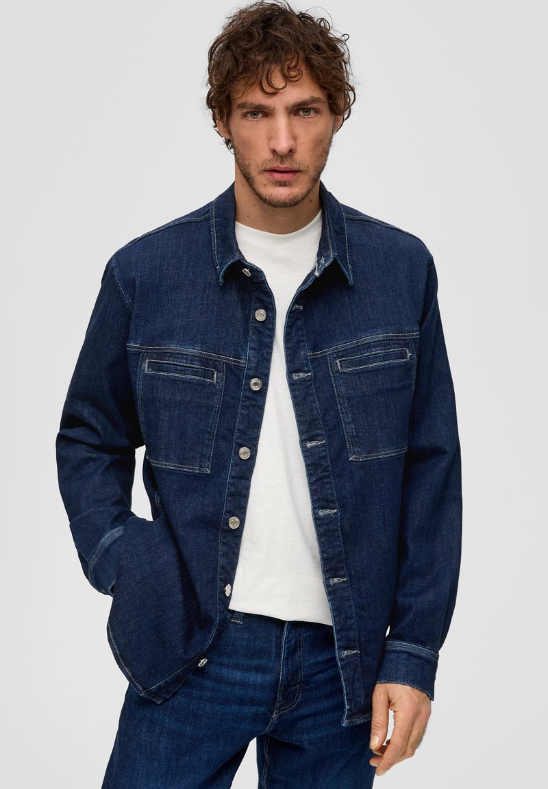 Donkerblauwe denimjack met een knoopsluiting aan de voorkant, twee borstzakken en contrasterende stiksels. Draagt over een wit t-shirt.