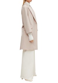Helles rosa maßgeschneidertes Kleid mit langen Ärmeln, offenem Vorderteil und Bindegürtel. Kombiniert mit weißen, hoch taillierten Hosen und einem weißen Oberteil. Angenehme Textur.