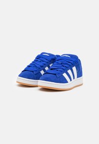 Μπλε σουέντ αθλητικά παπούτσια με λευκές ρίγες, στρογγυλή μύτη και μπλε κορδόνια. Η σόλα είναι λευκή με καφέ χρώμα από κάτω.