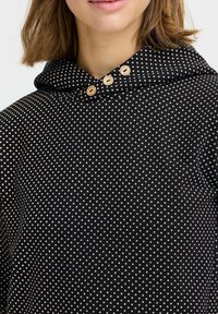 Felpa nera con pois bianchi, dotata di cappuccio e tre bottoni in legno decorativi sul collo. Tessuto morbido con una vestibilità comoda.