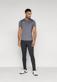 Γκρι αθλητικό t-shirt με κοντά μανίκια και λογότυπο Under Armour, σε συνδυασμό με σκούρο παντελόνι με πράσινες πλαϊνές ρίγες και λευκά αθλητικά παπούτσια.