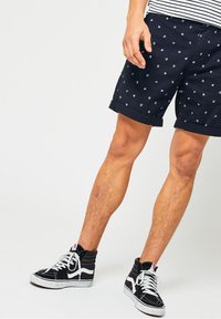 Námořnické šortky s bílými vzory kotvy, ohrnutými lemy a hladkým bavlněným materiálem. V kombinaci s černobílými sneakers s vysokým střelem.