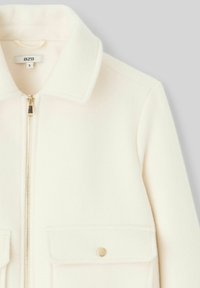 Veste en polaire crème avec un col, fermeture éclair dorée et deux poches avant avec fermetures à pression. Texture douce.