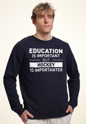 Mørkeblå sweatshirt med hvid tekst, der står "UDDANNELSE ER VIGTIG, MEN HOCKEY ER VIGTIGERE," med rund hals og lange ærmer.
