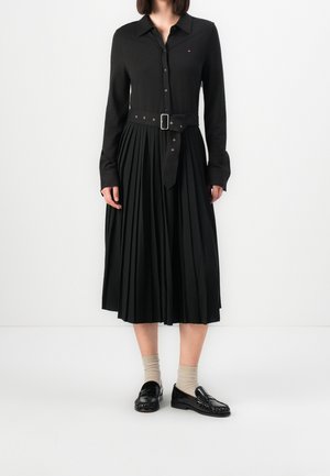 Femme portant une robe noire à manches longues avec col, jupe plissée et ceinture, chaussettes beiges et mocassins noirs, debout sur un fond blanc.