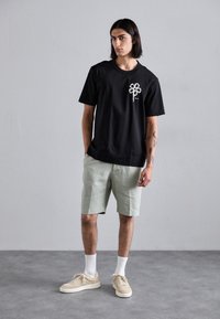 T-shirt en coton noir avec un graphique floral, associé à un short vert clair, des chaussettes blanches et des baskets beige sur un sol texturé.