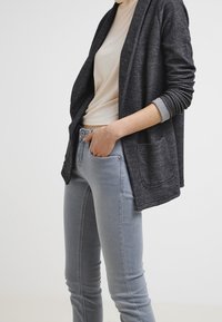 Tmavě šedý pletený cardigan s otevřeným předním dílem, dlouhými rukávy a předními kapsami, vrstvený přes světle béžový top a šedé skinny džíny.