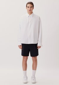 Camicia bianca con bottone, dal fit rilassato, maniche lunghe e colletto standard, abbinata a pantaloncini blu scuri e sneakers bianche.