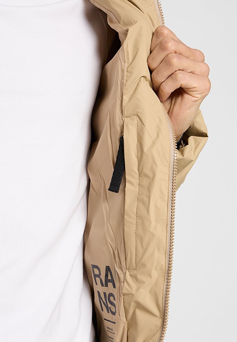 Une main tenant la fermeture éclair ouverte de la veste beige, révélant un tirette noire et le logo partiel "RAINS" à l'intérieur.