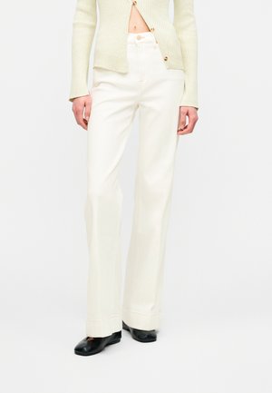 Osoba oblečená vo vysokým pásom bielych širších nohaviciach, svetlozelenom rebrovanom svetri na gombíky a čiernych plochých topánkach na jednoduchom pozadí.