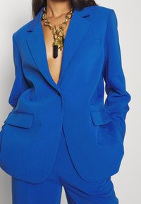 Woman wearing a bright blue tailored suit with hands in pockets and a chunky gold chain necklace with black pendant.