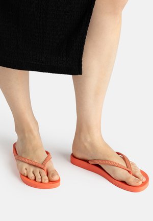 Füße, die leuchtend orangefarbene Flip-Flops mit strukturierten Riemen tragen, sichtbar vom Unterschenkel abwärts, stehend auf einer weißen Oberfläche mit einem schwarzen Rock.