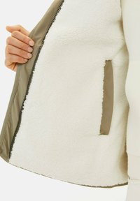 Fleecegefütterte Jacke mit einem weichen weißen Innenfutter, olivgrünem Außenmaterial und einer mit passendem Stoff vernähten Reißverschlusstasche.