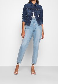 Mörkjeansjacka med knappar framtill och fickor, parat med ljusblå högmidjade jeans. Bärs med vita sandaler med remmar.