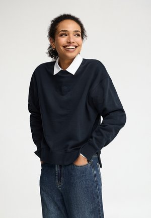Joven con cabello rizado sonriente, vestida con una sudadera negra sobre una camisa blanca con cuello y jeans azules, con las manos en los bolsillos.