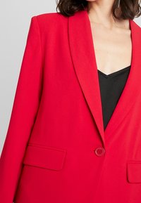 Roter Blazer mit Reverskragen, Ein-Knopf-Verschluss und zwei Fronttaschen. Hergestellt aus glattem Stoff mit strukturierter Passform. Schwarzes Oberteil darunter.