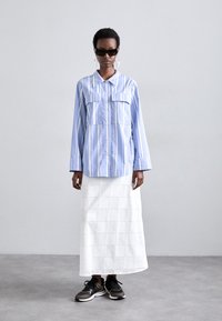 Light blue and white striped shirt with two front pockets, paired with a white eyelet maxi skirt and black athletic shoes. Dark sunglasses worn.
