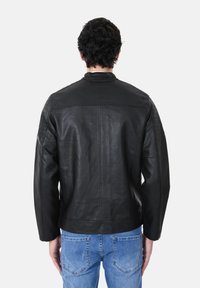 Giacca di pelle nera con una vestibilità leggermente ampia, caratterizzata da dettagli trapuntati sulle spalle, un colletto alto e una texture liscia. Abbinata a jeans blu.