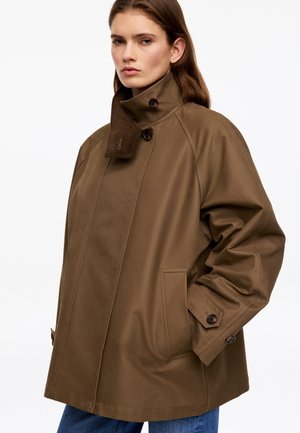 Femme portant un manteau marron oversize à col montant et boutons, posant les mains dans les poches devant un fond uni.