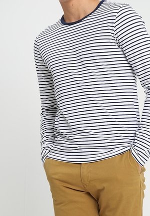 Personne portant une chemise blanche à manches longues avec de fines rayures horizontales noires et un pantalon marron moutarde, mains dans les poches.