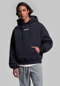 Hoodie bleu marine en tissu doux, avec une poche avant, des cordons de serrage ajustables et un petit logo blanc sur la poitrine.