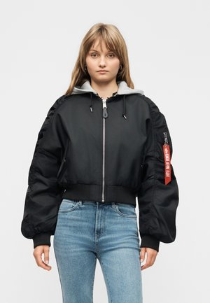 Fiatal nő fekete pufi dzsekiben, piros címkével az ujjon és kék farmernadrágban, egyszínű világos háttér előtt állva.