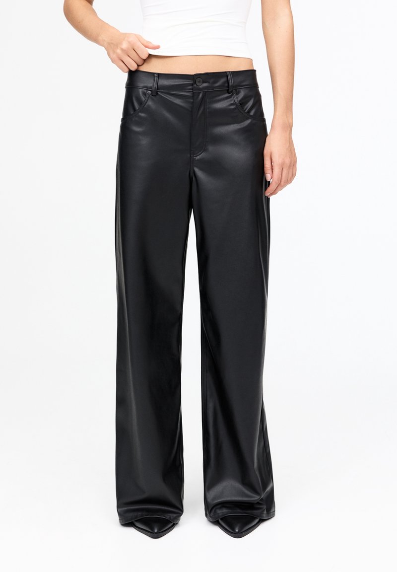 Femme portant un pantalon en cuir noir taille haute à jambes larges, associé à un débardeur blanc court, debout devant un fond blanc uni.